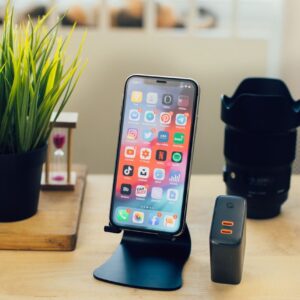 Arrangement of contemporary mobile phone on stand placed on wooden working table near uninterruptible power supply and photo camera lens
